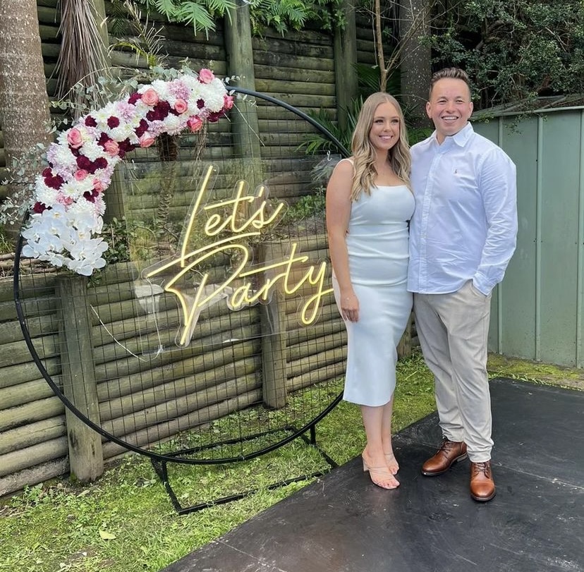 couple posing in front of a black mesh hoop backdrop with a lets party neon sign and florals